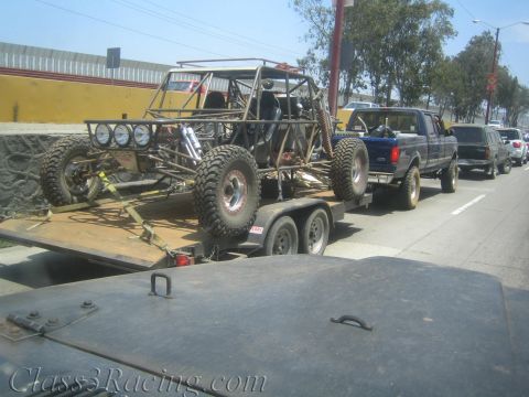 1990 Ford Bronco - tube-chassis off-road buggy rides on a flatbed trailer towed by a blue Ford pickup through a town street.
