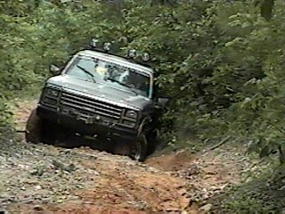 1980 Ford Bronco - This gray Ford Bronco is shown off-roading on a dirt path surrounded by trees. The SuperMotors logo is visible in the lower right corner.