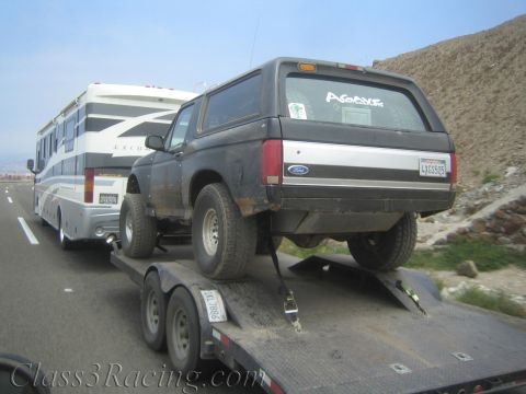 1990 Ford Bronco - muddy black Ford full-size SUV with a rear topper rides on a trailer towed behind a white motorhome.