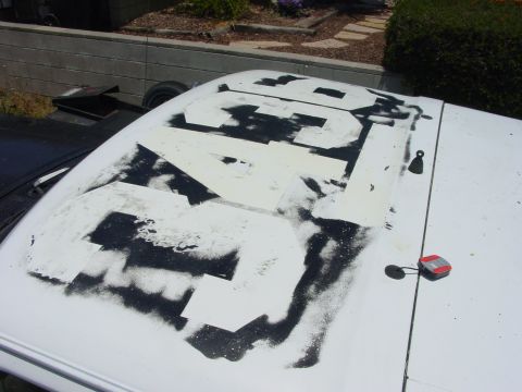 1994 Ford Bronco - Looking down on the white roof of an SUV where large black numbers are being spray-painted through cardboard stencils, with overspray still…