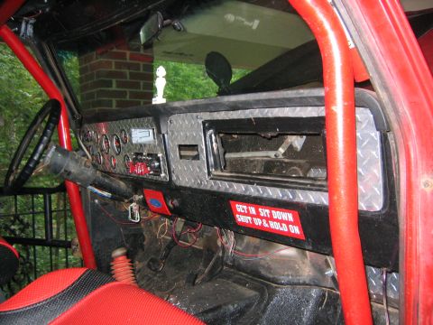 1979 Ford Bronco - The interior of a red vehicle features a diamond plate dashboard, a roll cage, and a humorous sticker on the dash.