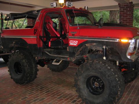 1979 Ford Bronco - Red lifted Ford Bronco off-road truck with roll cage and oversized mud tires parked under a brick carport.