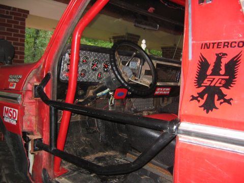 1979 Ford Bronco - Gutted red off-road truck interior showing roll cage, diamond-plate dash, aftermarket gauges and Interco STC eagle decal on door.