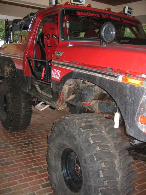 1979 Ford Bronco - Low-angle driver's-side view of a red lifted Ford Bronco on huge mud-terrain tires and black steel wheels, parked on a brick driveway under…