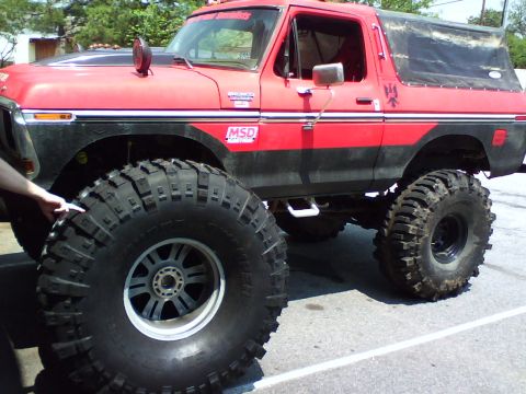1979 Ford Bronco - Low three-quarter front view of a lifted red and black full-size Ford Bronco on pavement with a black soft top, showing a loose oversized m…