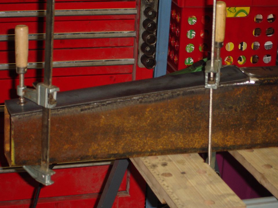 Rusted metal beam held by clamps on a workbench, with red tool chests in the background