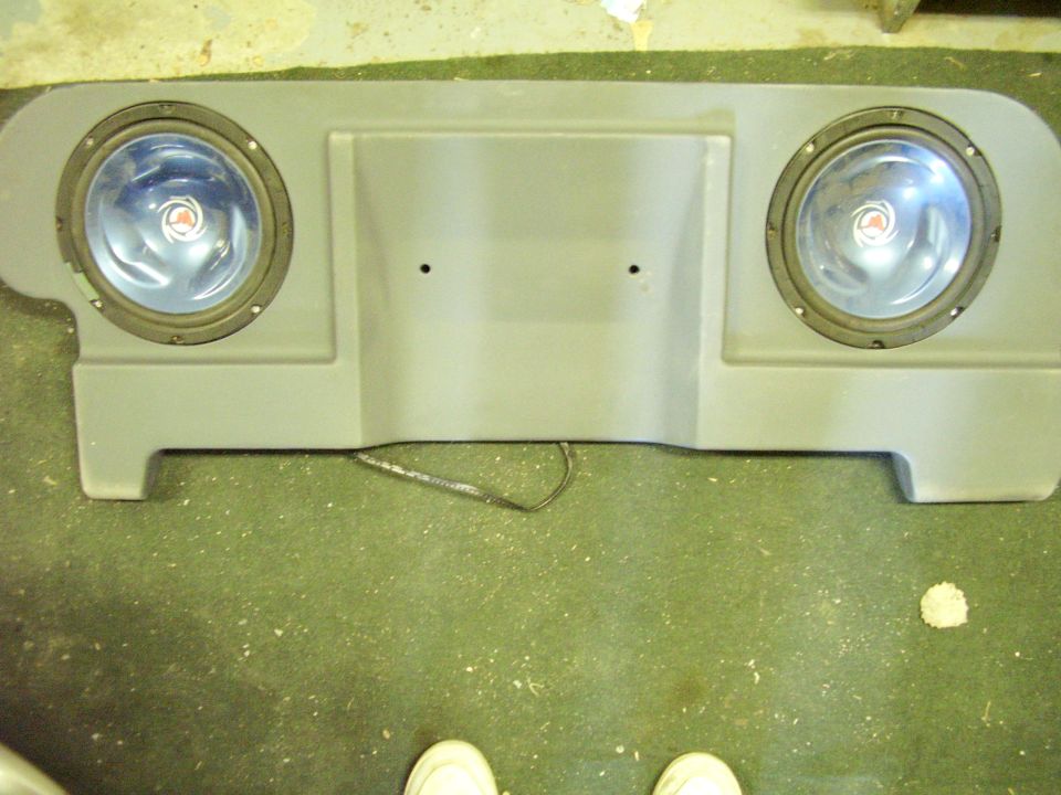 Gray speaker enclosure with two round speakers mounted, sitting on a green carpeted floor