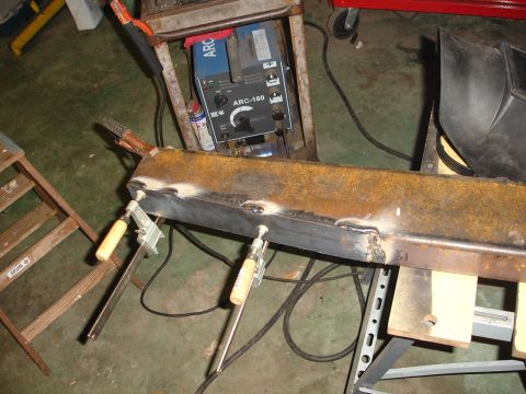 1989 Ford Bronco - A metal beam is being welded in a workshop setting, with a welding machine and clamps visible.