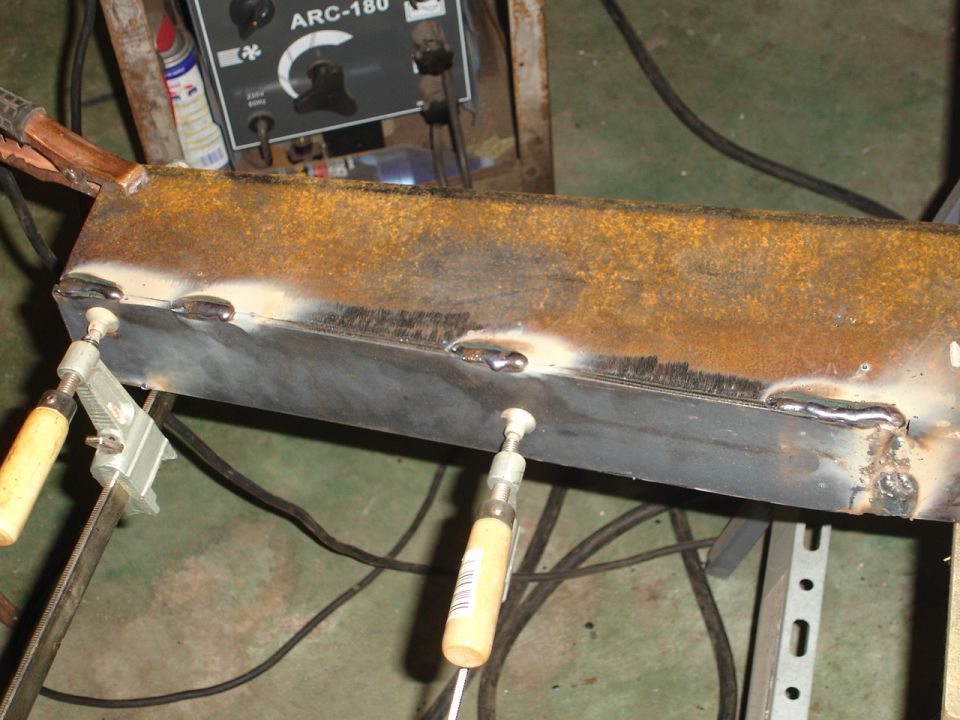 Rusty metal beam being welded, held by clamps, with a welding machine in background