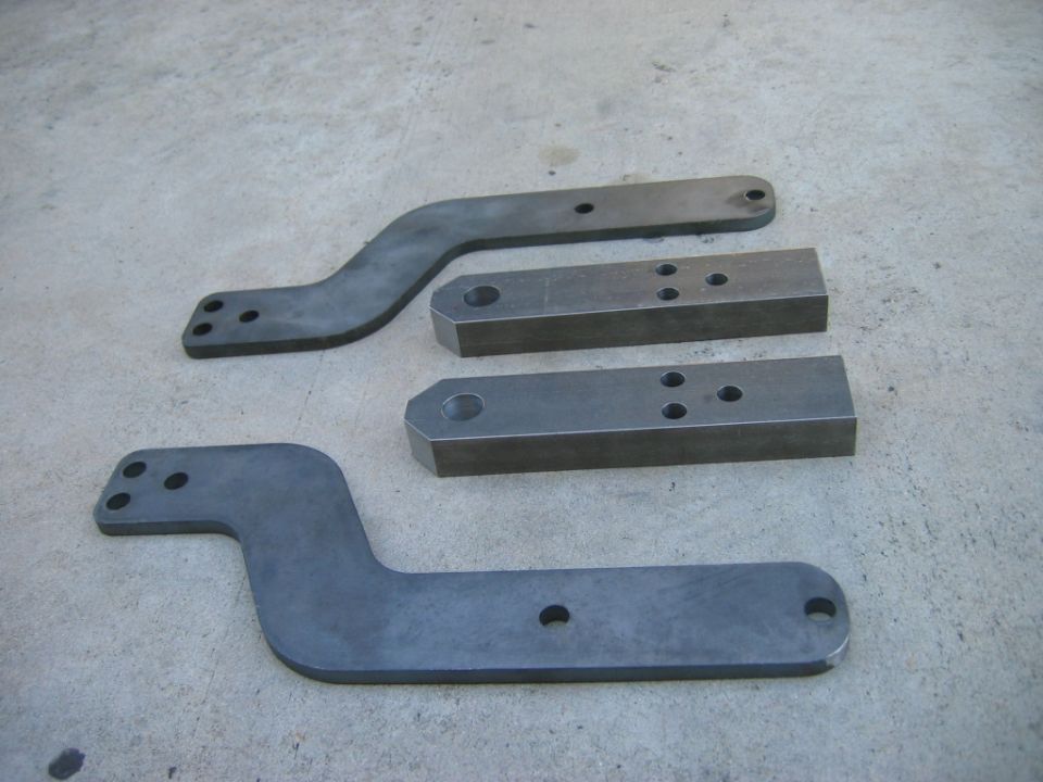 Four metal brackets with drilled holes on a concrete surface. Two are flat and curved, two are rectangular blocks