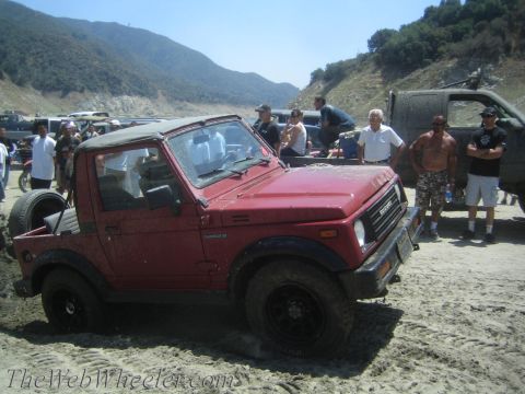 1987 Suzuki Samurai - A group of people gather around a red Suzuki Samurai as it sits on a sandy, off-road area.
