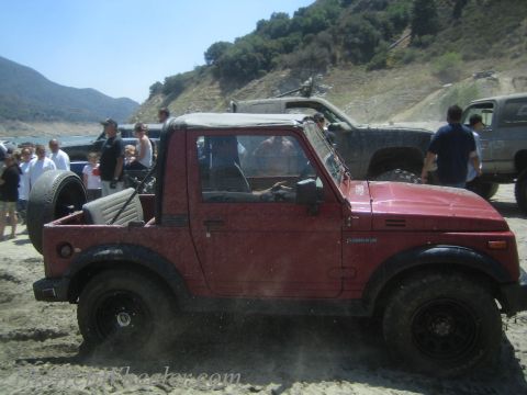 1987 Suzuki Samurai - A red 1987 Suzuki Samurai is driven off-road with other vehicles and people in the background.