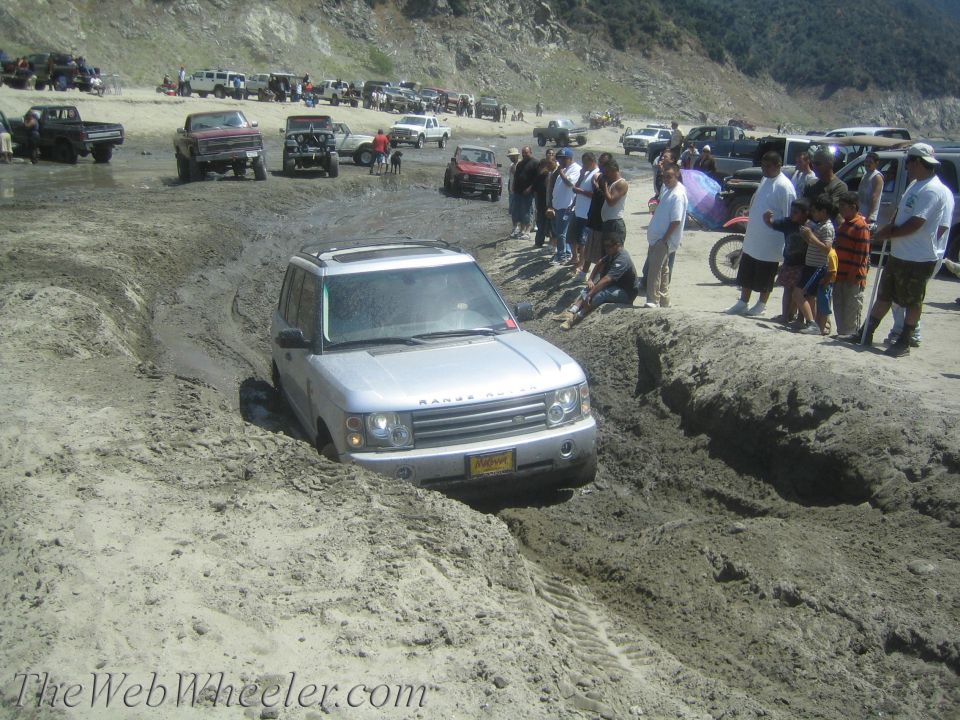 Silver Range Rover stuck in a deep mud trench, with spectators watching. Other vehicles are parked in the background