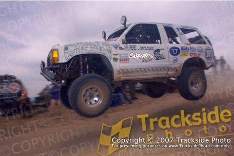 1992 Ford Explorer - A modified SUV is captured mid-air during a race, kicking up dust on a sandy track.