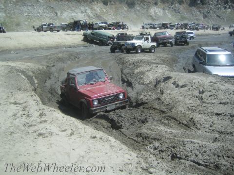 1987 Suzuki Samurai - A red 1987 Suzuki Samurai tackles a challenging muddy terrain, with other off-road vehicles visible in the background.