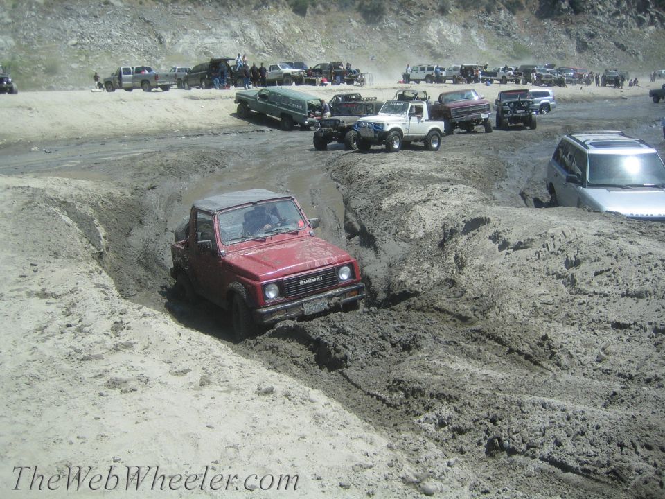 Red 1987 Suzuki Samurai navigating a muddy ditch with its top down