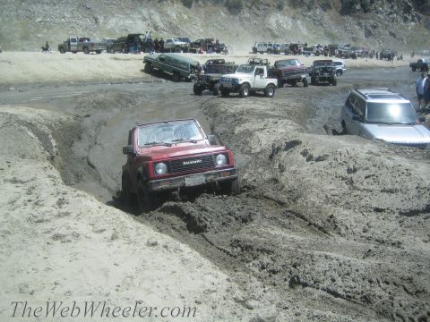 1987 Suzuki Samurai - A red 1987 Suzuki Samurai tackles a challenging muddy terrain, with other vehicles visible in the background.