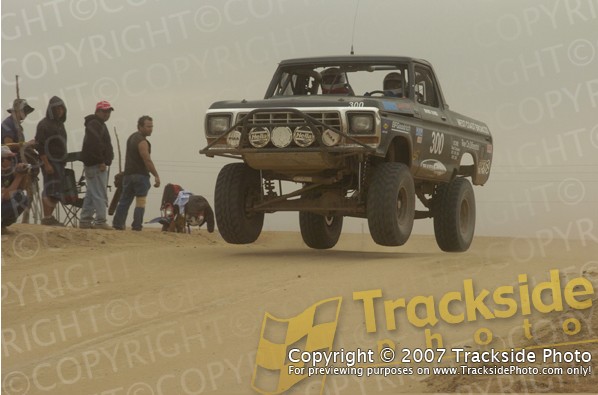 Off-road truck mid-air jump, number 300 on door, people watching on sandy dune