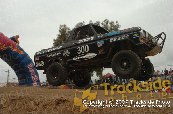 Off-road racing truck with number 300 airborne over a dirt jump, Red Bull arch in background
