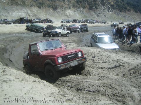1987 Suzuki Samurai - A red 1987 Suzuki Samurai tackles a challenging muddy terrain with other vehicles and spectators in the background.