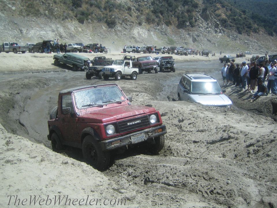 Red 1987 Suzuki Samurai stuck in mud pit with other vehicles and people watching