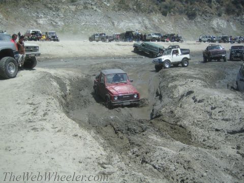 1987 Suzuki Samurai - Several off-road vehicles are gathered at a muddy terrain, with one red Suzuki Samurai navigating a deep mud trench.