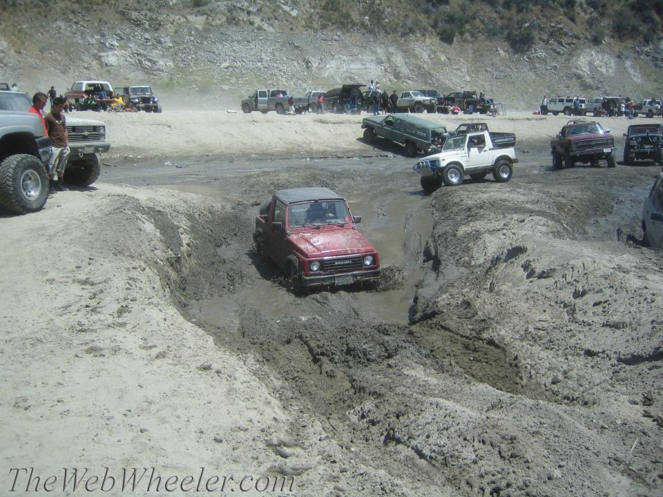 Red 1987 Suzuki Samurai driving through a mud pit with other off-road vehicles around