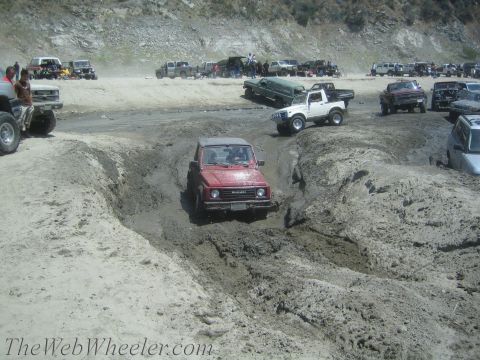 1987 Suzuki Samurai - A red Suzuki Samurai attempts to traverse a challenging muddy terrain with other off-road vehicles present.