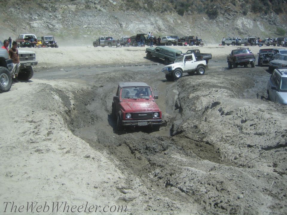 Red Suzuki Samurai navigating a muddy rutted track, front view. The vehicle has a soft top and appears to be from the late 1980s