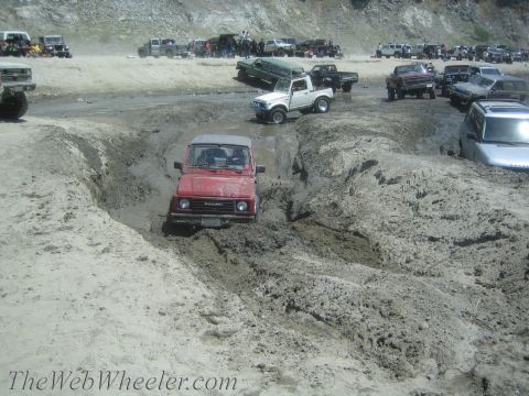 1987 Suzuki Samurai - A red 1987 Suzuki Samurai navigates a challenging muddy terrain, with other vehicles visible in the background.