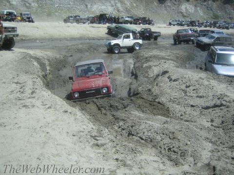1987 Suzuki Samurai - Off-roaders navigate a muddy track, with a red Suzuki Samurai prominently stuck in the mire.