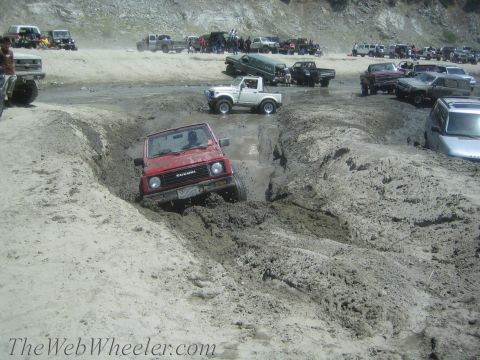1987 Suzuki Samurai - Several off-road vehicles are gathered on a sandy terrain, with one red Suzuki Samurai navigating a challenging muddy path.