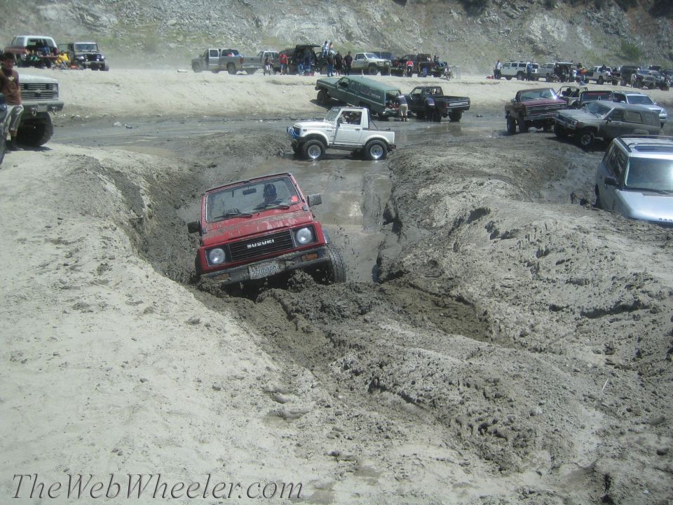 Red Suzuki Samurai stuck in a muddy ditch, with other vehicles in the background on a sandy area