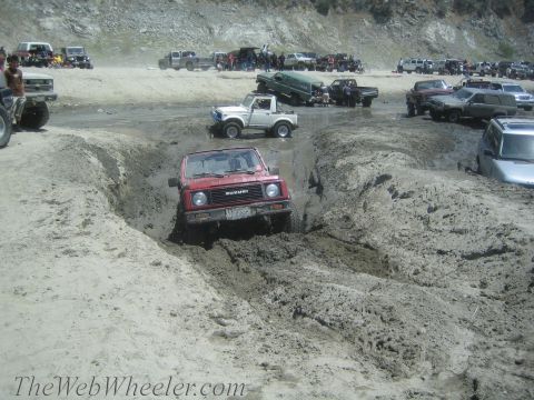 1987 Suzuki Samurai - Multiple off-road vehicles are seen in a muddy terrain, with a red Suzuki Samurai attempting to traverse a deep rut.