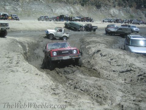 1987 Suzuki Samurai - A red 1987 Suzuki Samurai tackles a challenging muddy terrain, with other vehicles visible in the background.