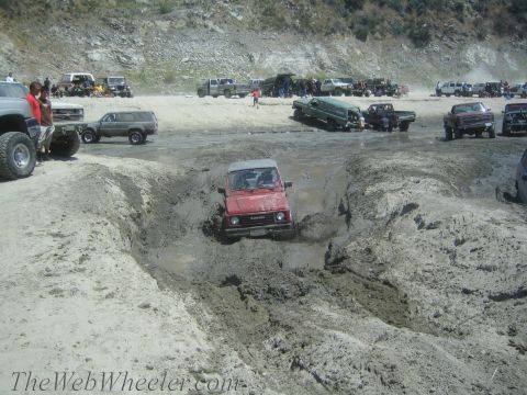 1987 Suzuki Samurai - A red 1987 Suzuki Samurai tackles a challenging muddy obstacle course with other vehicles in the background.
