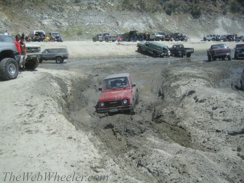 1987 Suzuki Samurai - A red 1987 Suzuki Samurai tackles a challenging muddy obstacle course with other vehicles in the background.