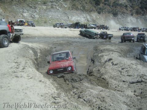 1987 Suzuki Samurai - Multiple off-road vehicles navigate a challenging muddy terrain with a red Suzuki Samurai prominently stuck in a ditch.