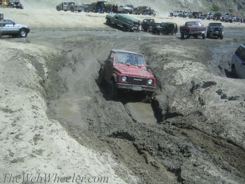 1987 Suzuki Samurai - A red Suzuki Samurai tackles a challenging mud pit during an off-road event, with spectators and other vehicles nearby.
