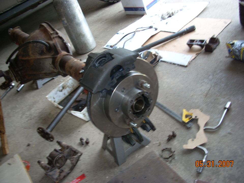 Vehicle rear axle assembly with brake rotor and caliper on jack stands. Tools and other parts are scattered around