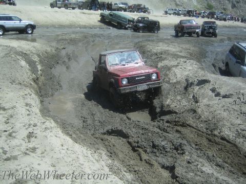 1987 Suzuki Samurai - A red 1987 Suzuki Samurai navigates a challenging muddy terrain, with other vehicles visible in the background.