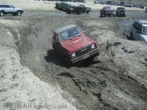 1987 Suzuki Samurai - A red 1987 Suzuki Samurai tackles a challenging muddy off-road obstacle with other vehicles visible in the background.