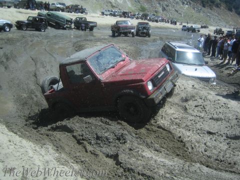 1987 Suzuki Samurai - A red Suzuki Samurai navigates a muddy off-road course, with spectators and other vehicles nearby.