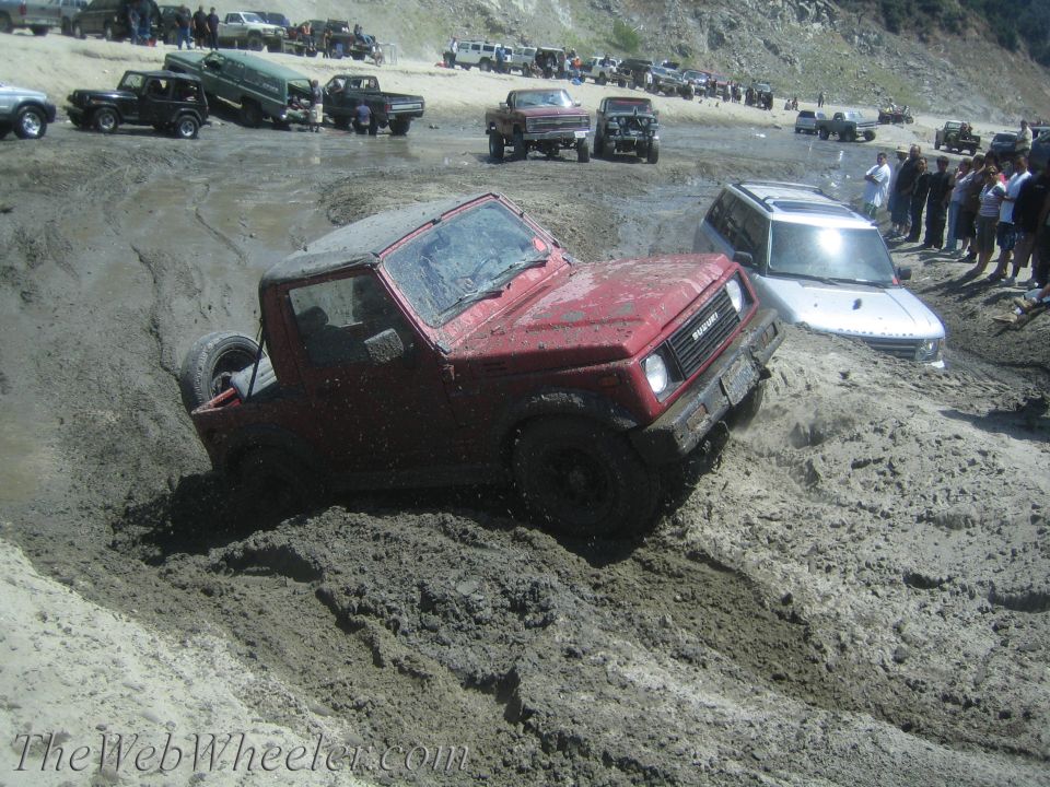 Red Suzuki Samurai stuck in mud, with a silver SUV partially submerged behind it. Other vehicles and people are visible on a muddy bank in the background