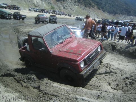 1987 Suzuki Samurai - A red Suzuki Samurai struggles in thick mud, with spectators gathered on the bank behind it.