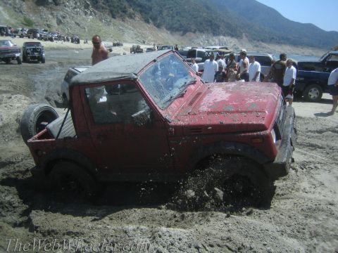 1987 Suzuki Samurai - A red 1987 Suzuki Samurai is stuck in thick mud, with other vehicles and people in the background at a lake or reservoir.