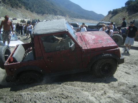 1987 Suzuki Samurai - Red Suzuki Samurai struggling in mud with a crowd gathered around.
