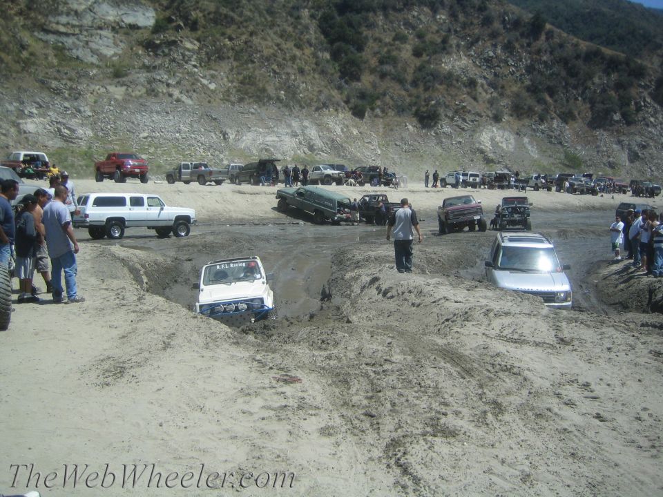 White Suzuki Samurai stuck in mud pit, front wheels submerged. "A.P.L. Racing" visible on windshield