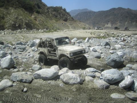 1987 Suzuki Samurai - A tan 1987 Suzuki Samurai navigates a rocky riverbed with mountains in the background.