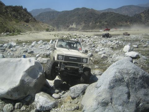 1987 Suzuki Samurai - A white 1987 Suzuki Samurai tackles a challenging rocky course in a dry riverbed, with other vehicles visible in the background.